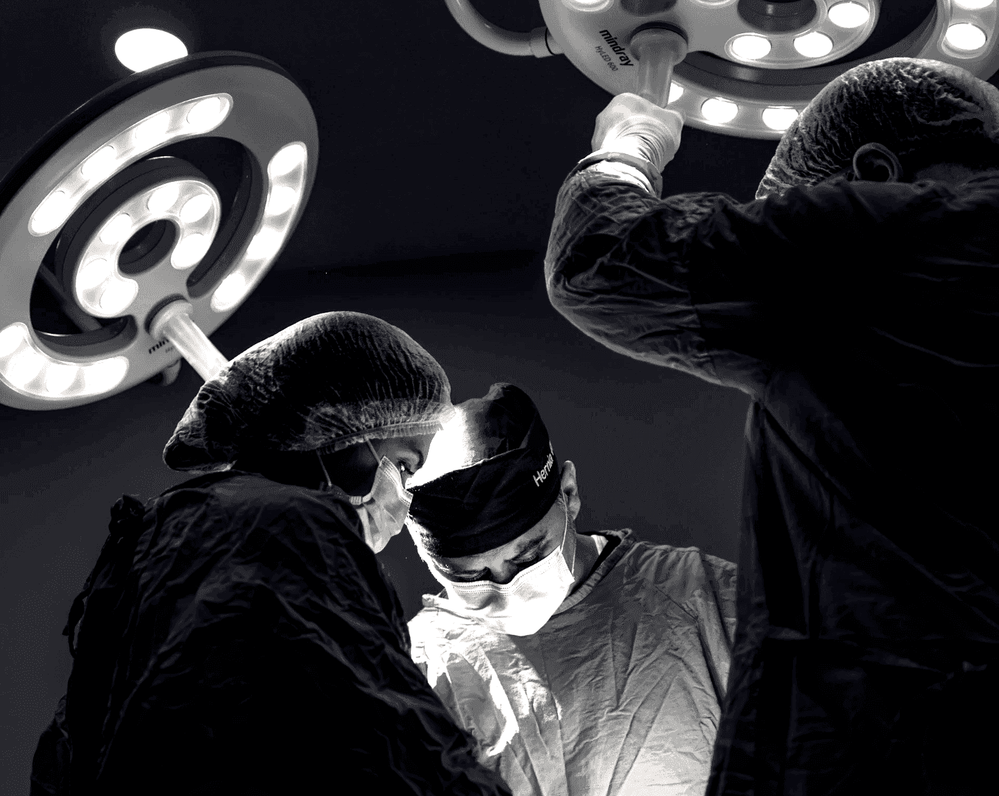 Black and white photo of surgeons in masks and gowns focused on a procedure under bright overhead operating lights